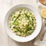 Creamy spring vegetable risotto with asparagus, peas, and zucchini topped with shaved Parmesan and lemon zest in a white bowl